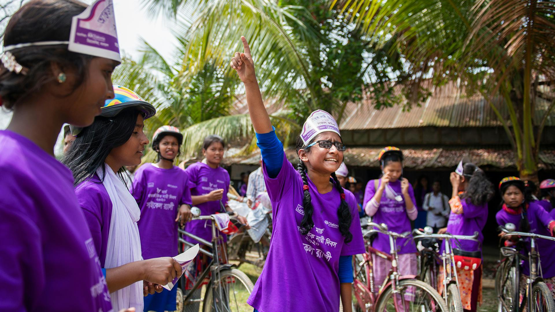 En flicka står mitt i en grupp av andra flickor med cyklar och pratar och gestikulerar med handen. Alla flickor har likadana lila t-shirts på sig.