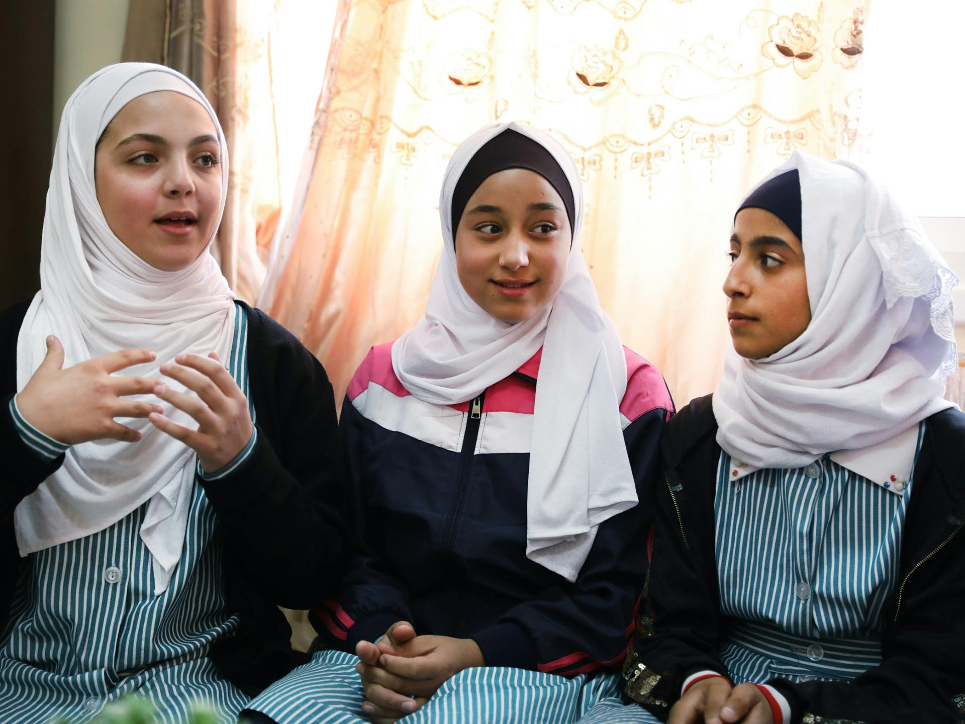 Tre unga flickor i hijab pratar och gestikulerar. Bakom dem syns ett fönster med en spetsgardin.