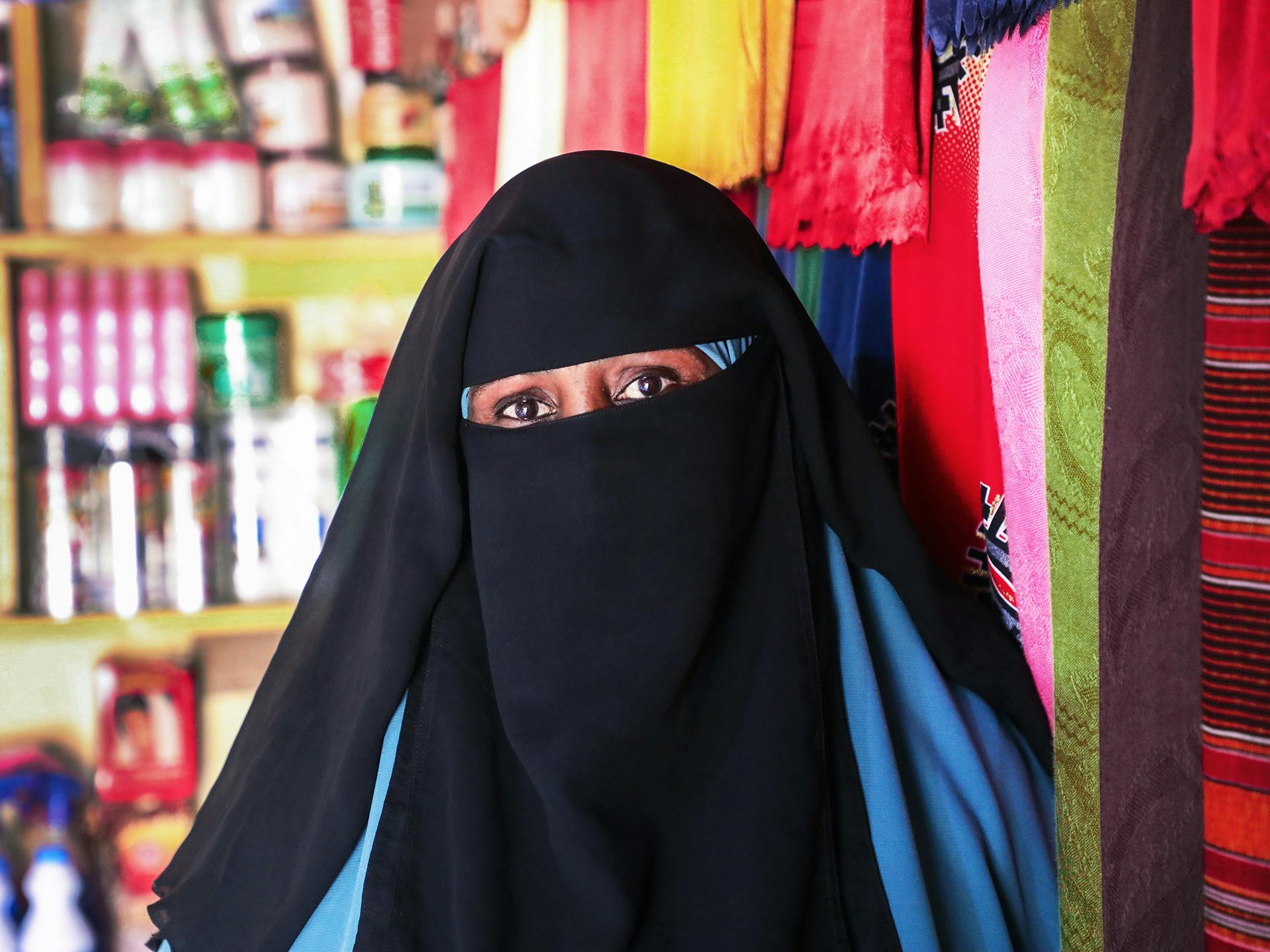 Närbild av kvinna i svart niqab och blå hiijab i en butik. I bakgrunden syns hyllor med livsmedelsprodukter och tyger i olika färger och mönster.