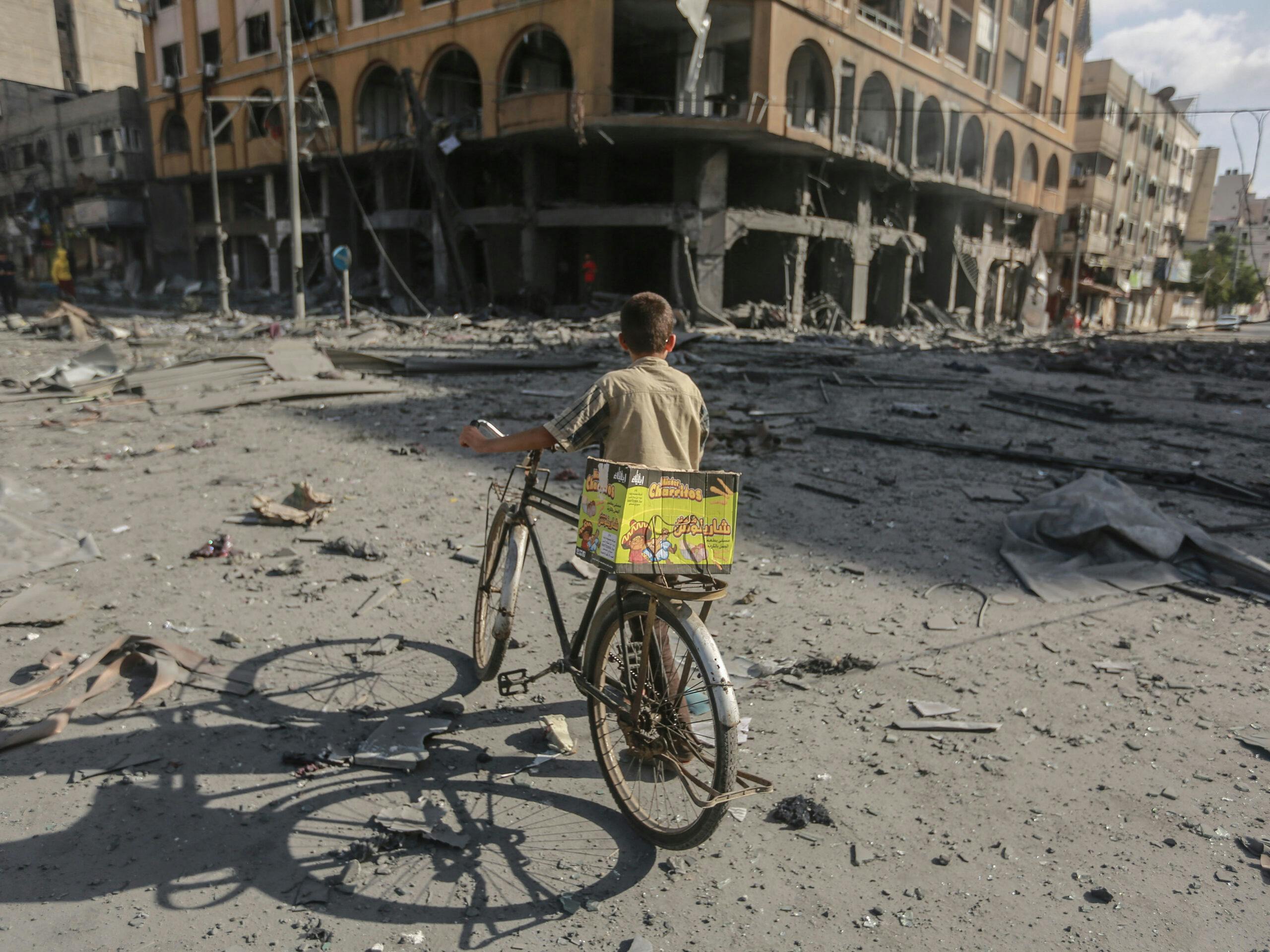 Ett barn står med en cykel och ryggen mot kameran, och ser mot ett sönderbombat hus.