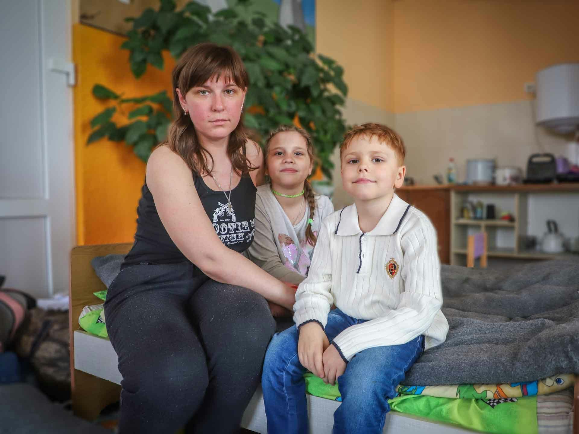 En kvinna och två barn sitter på en säng och tittar in i kameran
