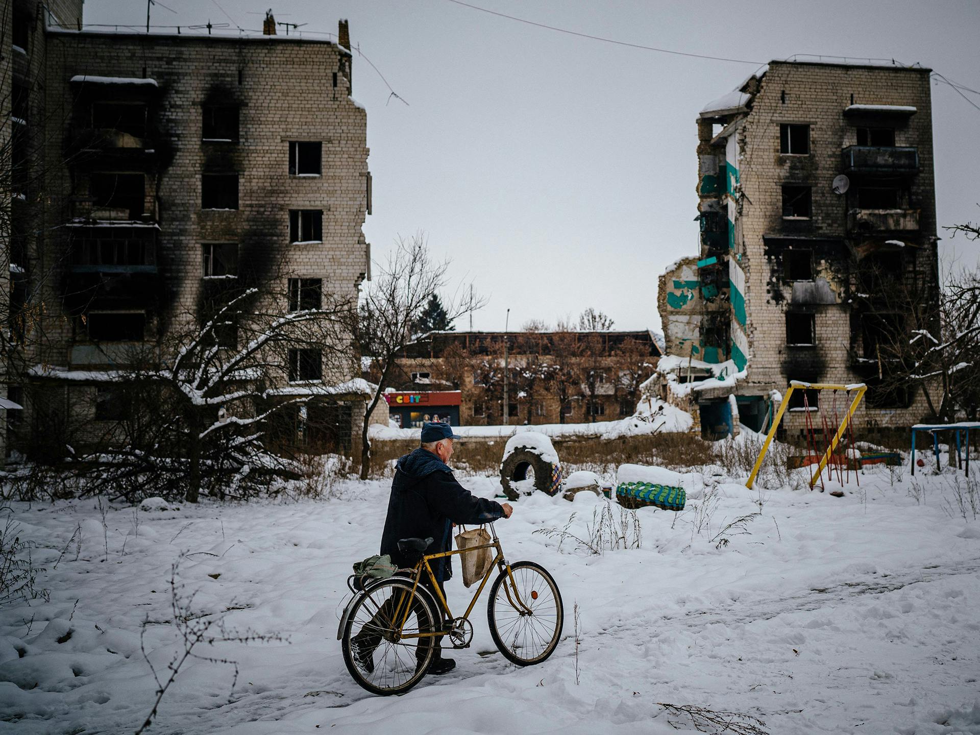 En man leder en cykel över snöiga gator. I bakgrunden syns förstörda hus.