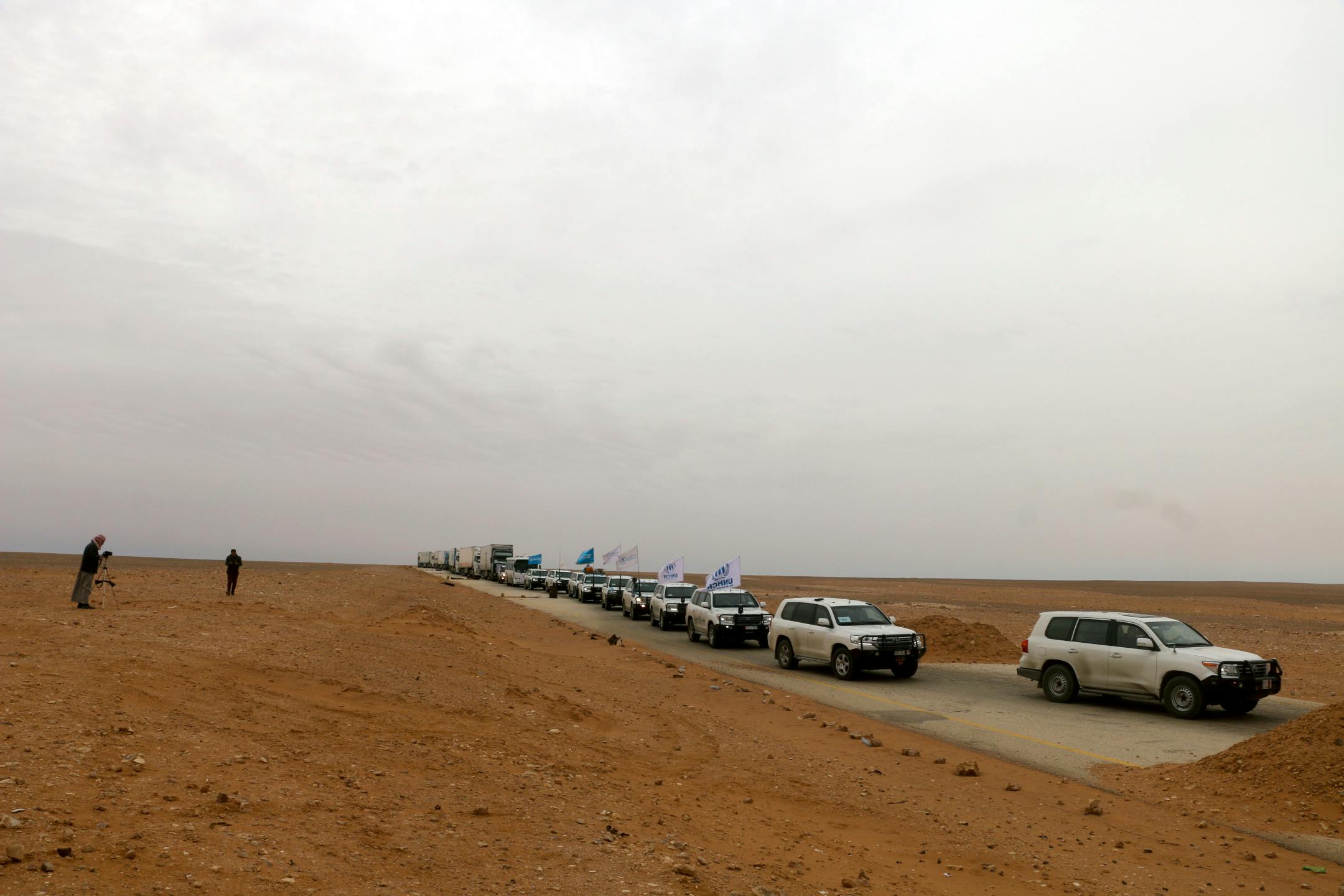 A convoy of cars with United Nations flags driving through dry land.