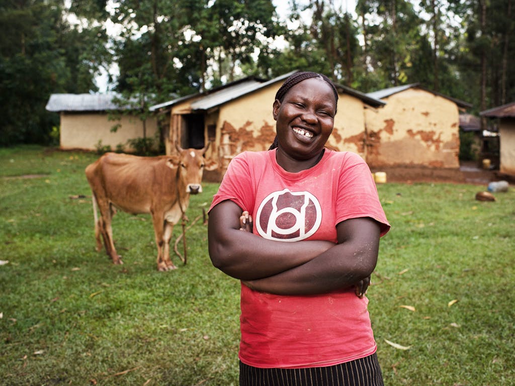 A smiling woman with her arms crossed on her chest. In the background there is a cow and a house.