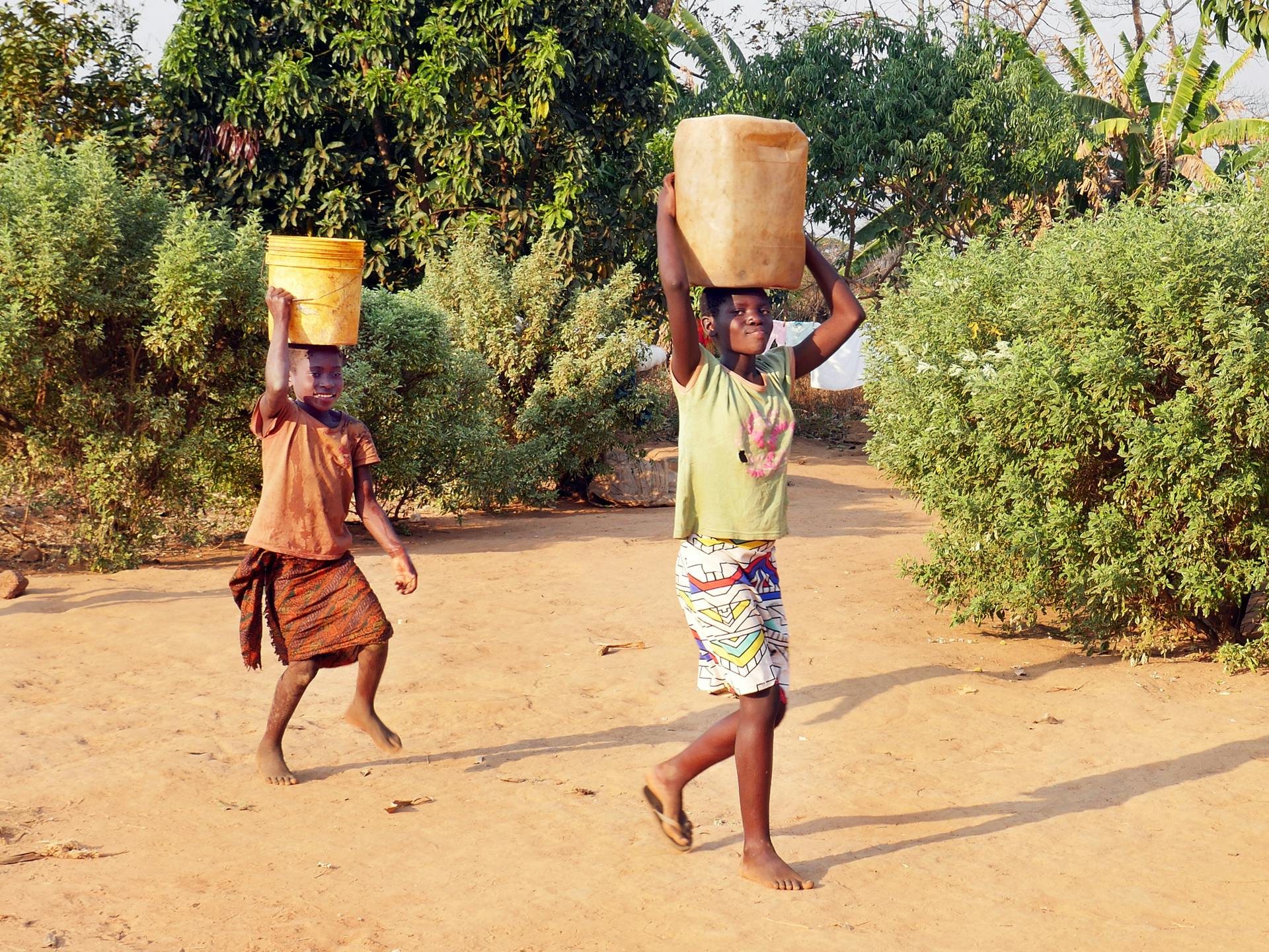 Two Zambian children carrying water containers on their heads.